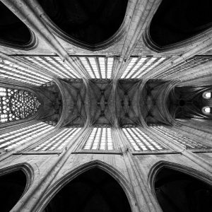 Photographie en noir et blanc d'une voûte d'église vue en contre-plongée, avec ses nervures de pierre et ses ouvertures lumineuses.