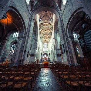 Allée centrale d’une église bordée de chaises, sous de hautes voûtes, en direction du chœur lumineux.