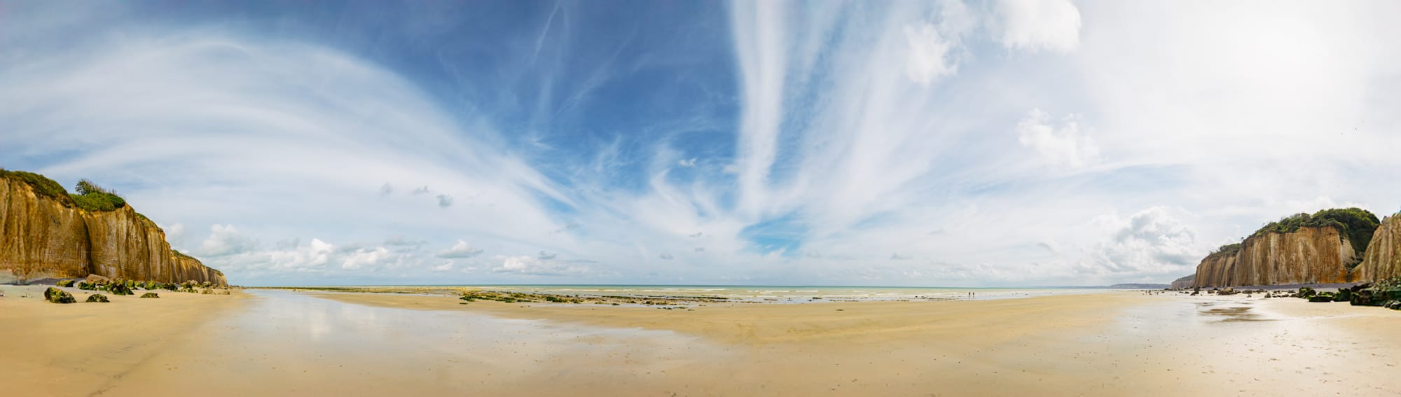Panorama d’une plage ouverte sur la mer entre deux falaises, sous un ciel parcouru de nuages.