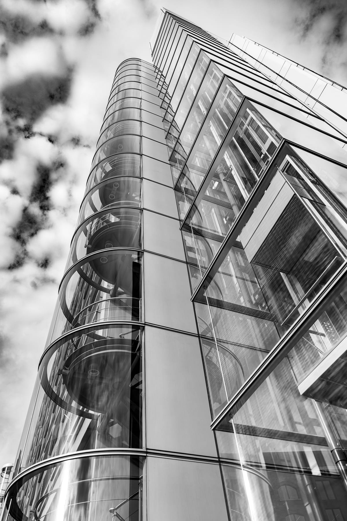 Vue en contre-plongée d'une cage d'escalier vitrée et des lignes verticales d'un bâtiment moderne se détachant sur le ciel.