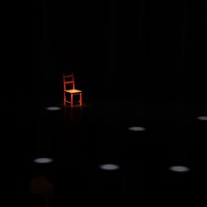 Une chaise rouge éclairée, isolée dans un vaste espace sombre ponctué de quelques taches de lumière.