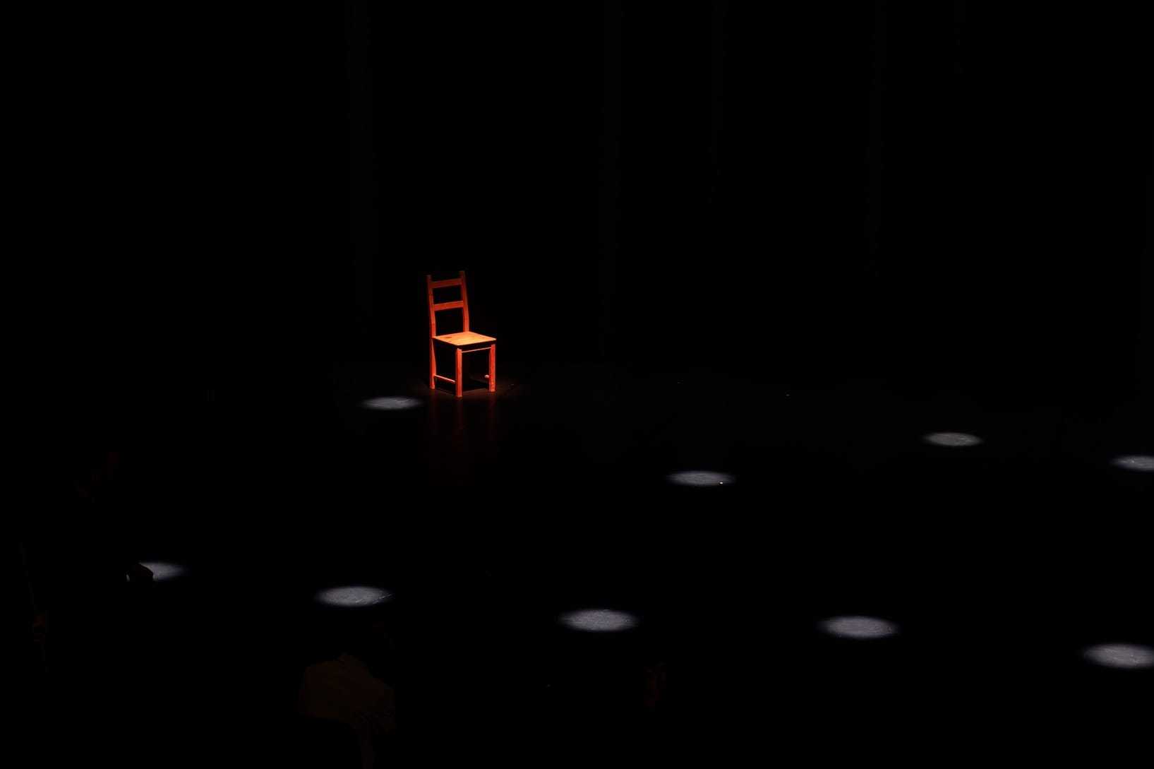 Une chaise rouge éclairée, isolée dans un vaste espace sombre ponctué de quelques taches de lumière.